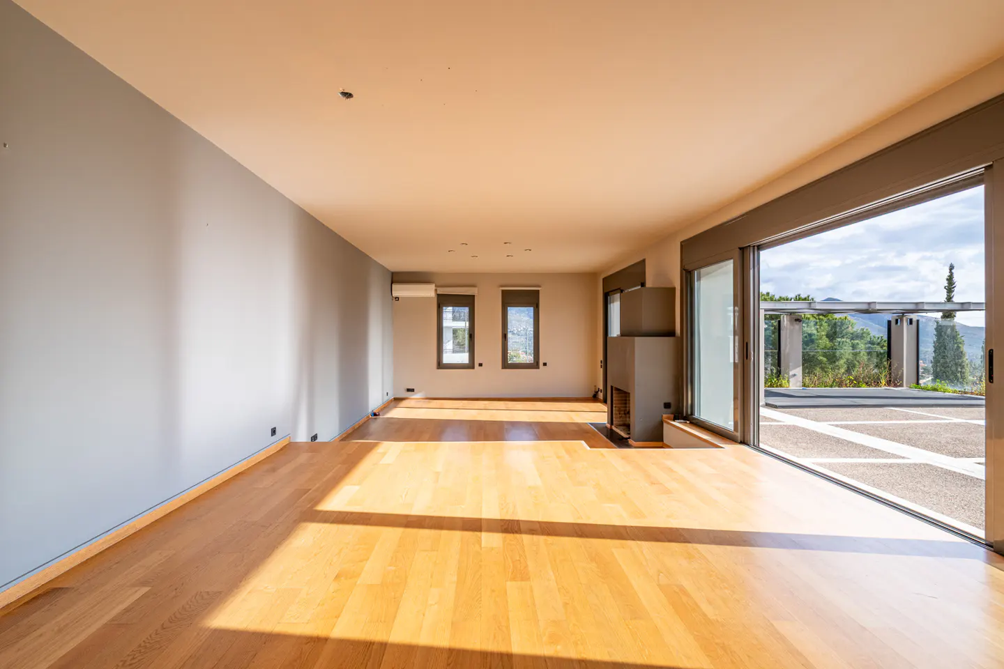 Bright, empty room with hardwood floors, gray walls, and a modern fireplace. Sliding glass doors open to a patio with a view of trees and mountains.