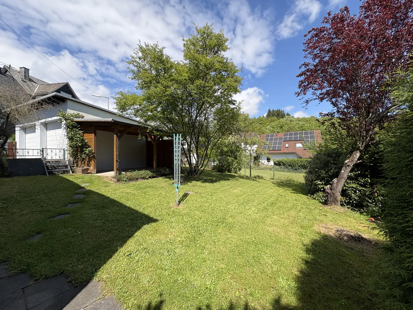 A lush green lawn with trees, a white house with a covered patio, and a clothesline under a partly cloudy sky.