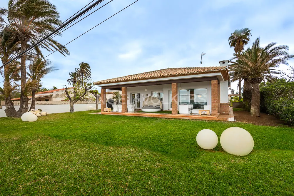 Exterior of a white, single-story house with a red tile roof and a covered porch with outdoor furniture, surrounded by a green lawn and palm trees.