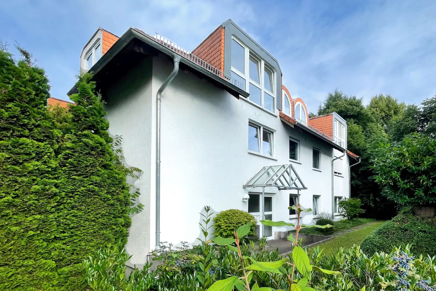 Exterior view of a white two-story house with a red tile roof and a glass-covered entrance. Green trees surround the house.