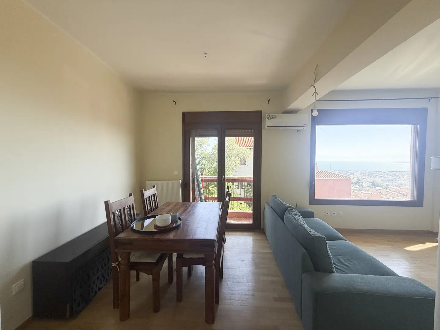Bright living room with a wooden dining set, blue sofa, and large window offering a scenic view of the city and sea.