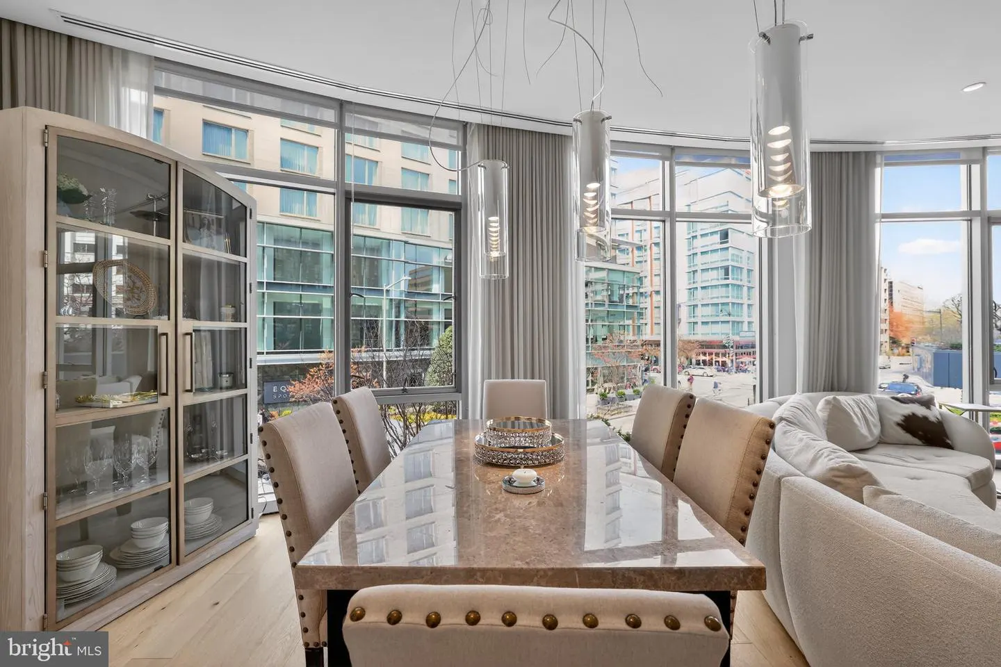 Bright dining room with a marble table, upholstered chairs, and a glass cabinet. Large windows offer a city view. Cylindrical pendant lights hang above.