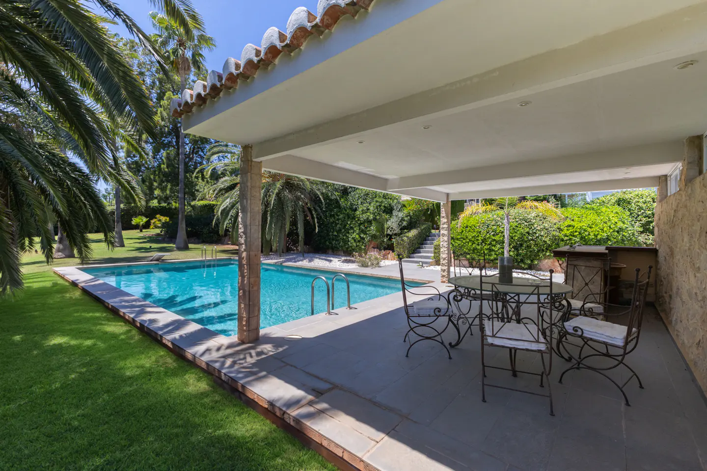 Outdoor patio with a pool. Table and chairs under a covered area. Palm trees and green lawn surround the pool.