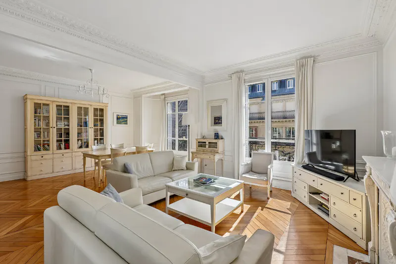 Bright living room with white sofas, glass coffee table, wooden floors, and large windows overlooking a city street.