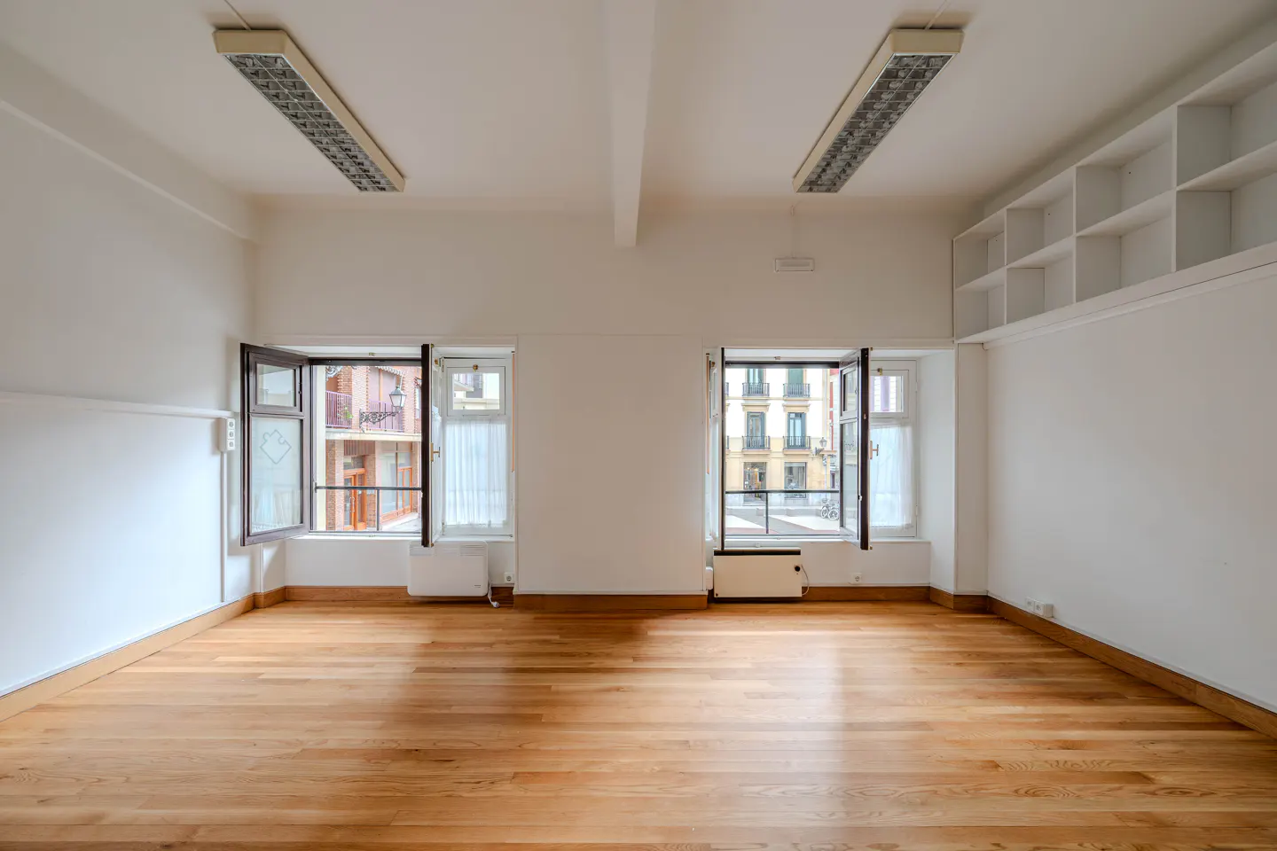Empty room with hardwood floors, white walls, and two open windows showing city buildings. Two fluorescent lights on the ceiling.