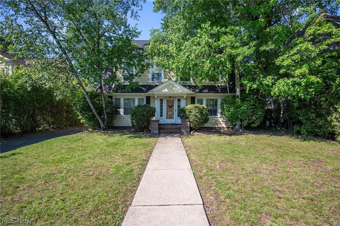 Two-story yellow house with a gray walkway leading to the front door, surrounded by green trees and lawn.