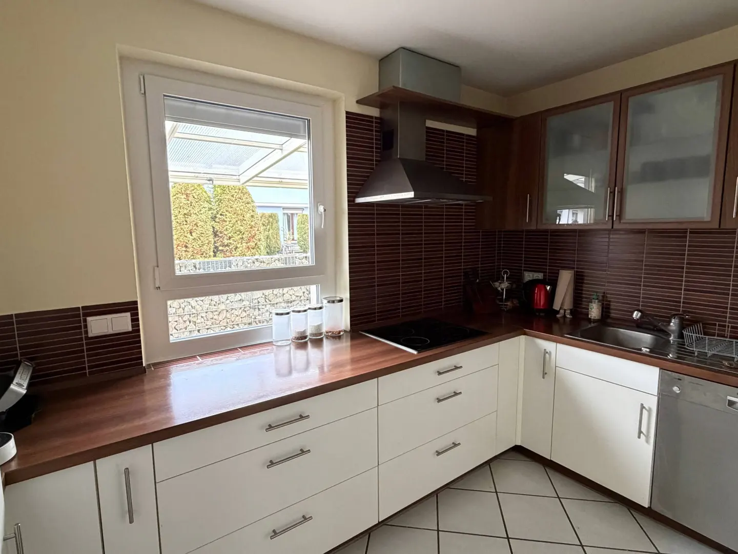 A kitchen with white cabinets, brown countertops, and a window overlooking a garden. The backsplash is brown tile.