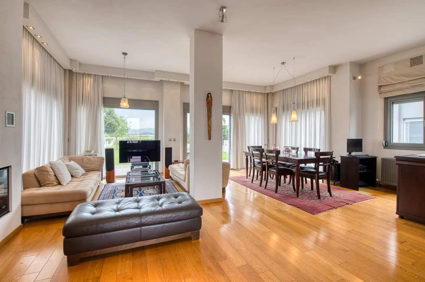 Bright, open-concept living and dining area with hardwood floors, tan sofas, and a dark wood dining table set on a red rug. Large windows with white curtains.