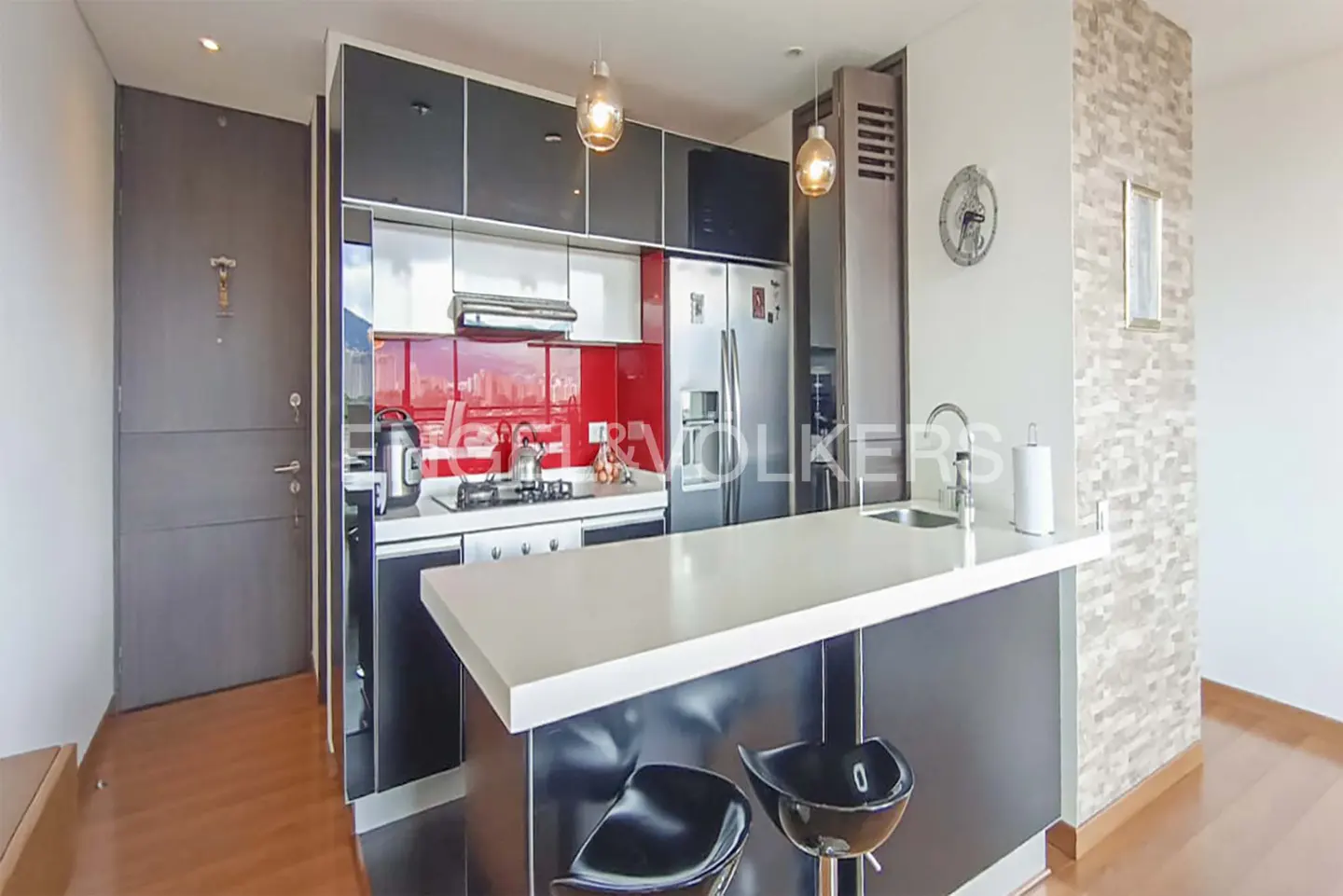 A modern kitchen with black cabinets, a white countertop island with two black stools, and stainless steel appliances. A red backsplash adds a pop of color.