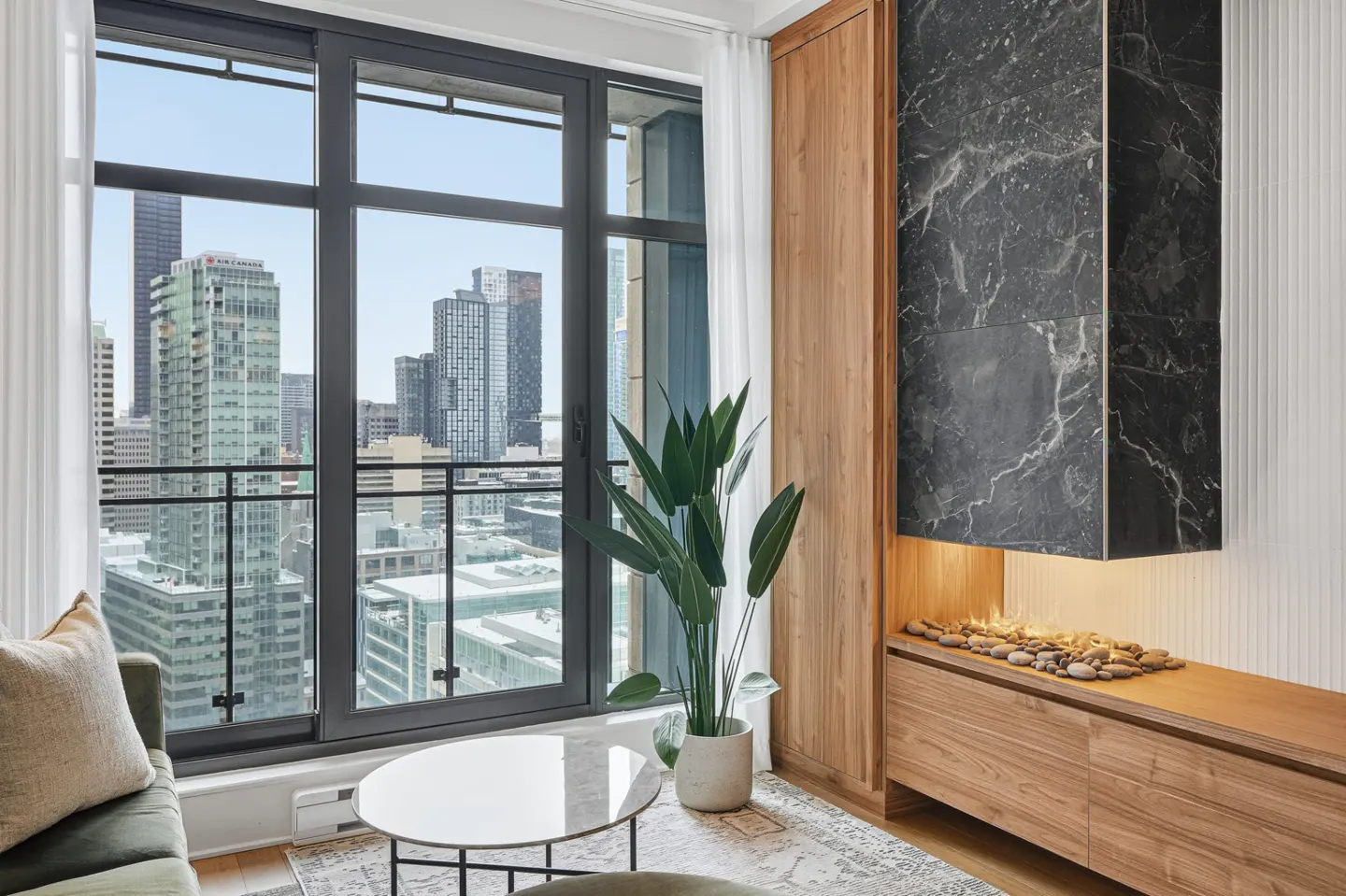 Living room with a city view. A black marble fireplace with pebbles sits next to a large window with sheer curtains.