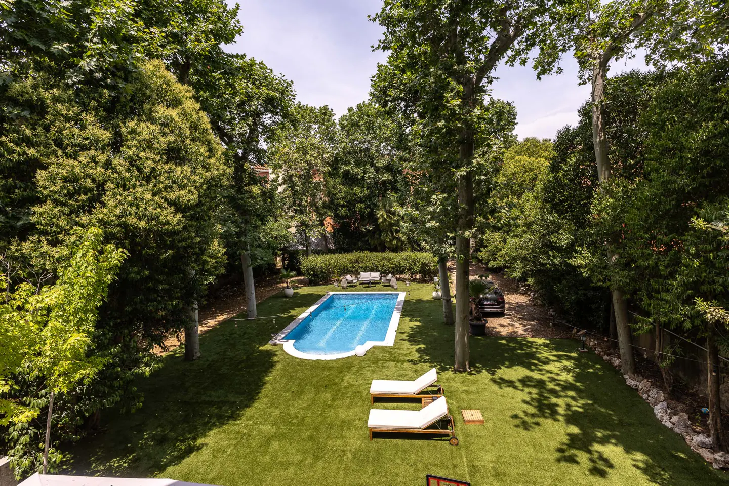 A lush backyard with a blue swimming pool, two lounge chairs on a green lawn, and tall trees surrounding the area.
