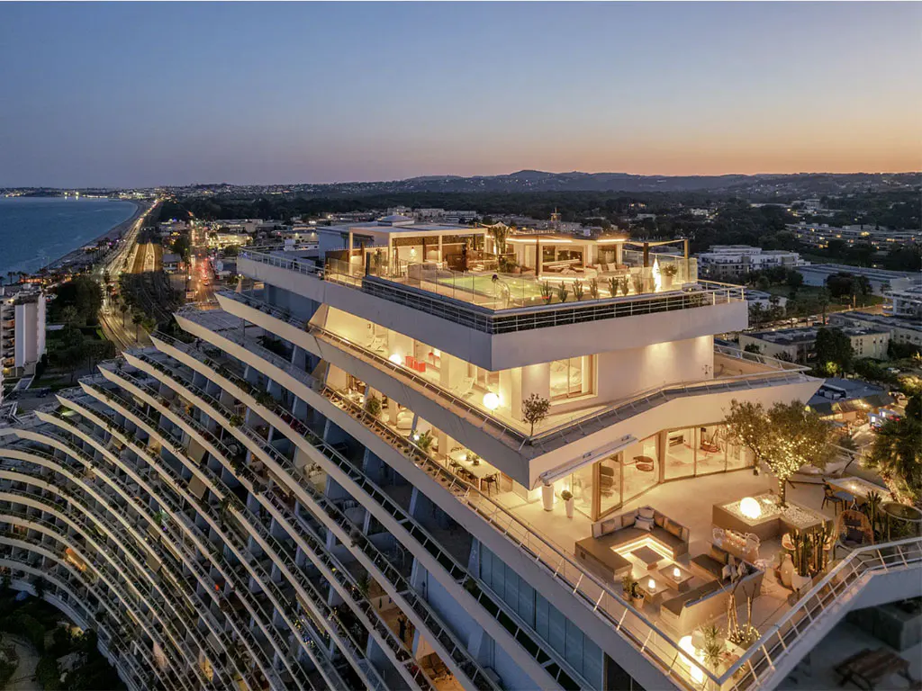 Aerial view of a modern, white, terraced building with rooftop pool and lounge areas at dusk, overlooking the coastline.