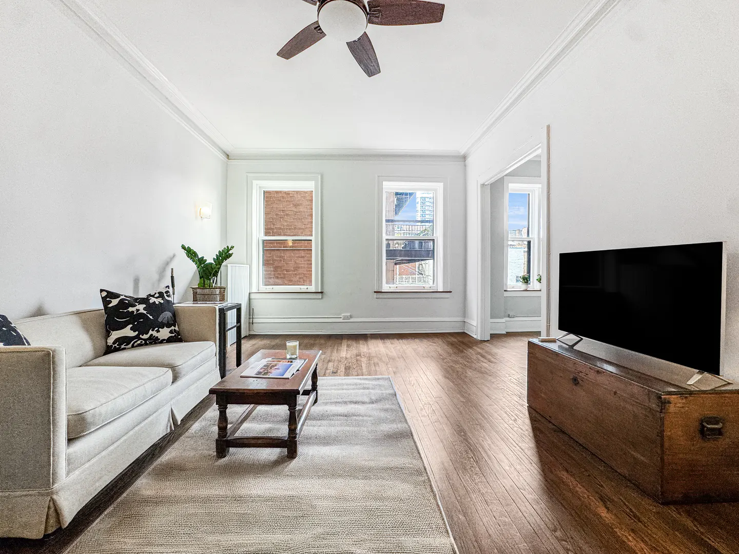 Living room with white walls, hardwood floors, and a ceiling fan. A beige sofa, wood coffee table, and TV on a trunk are visible. Windows offer a city view.