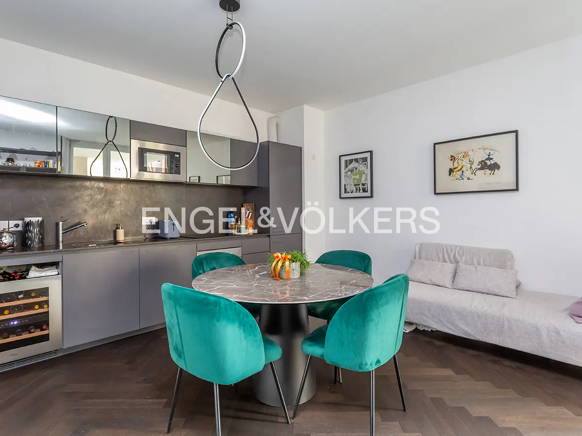 A modern apartment interior with a gray kitchen, round marble table, and four teal velvet chairs. A light gray sofa sits against a white wall with framed art.