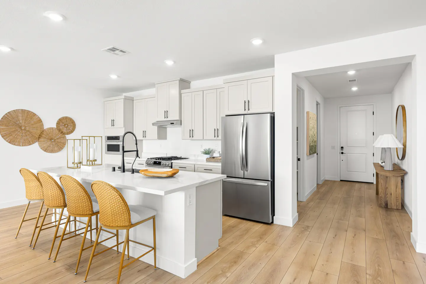 Bright, modern kitchen with white cabinets, stainless steel appliances, and a white quartz island with four yellow woven bar stools. Wood floors lead to a hallway.