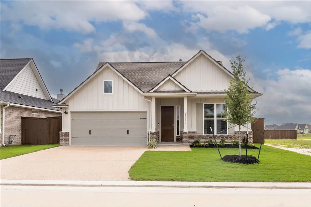 A single-story home with light siding, a gray garage door, and a brown front door. A green lawn and cloudy sky complete the scene.