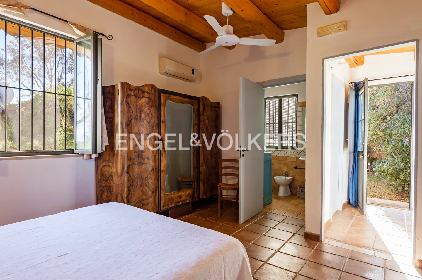 Bedroom with white bedspread, wood ceiling beams, and terracotta tile floor. A bathroom and outdoor patio are visible.