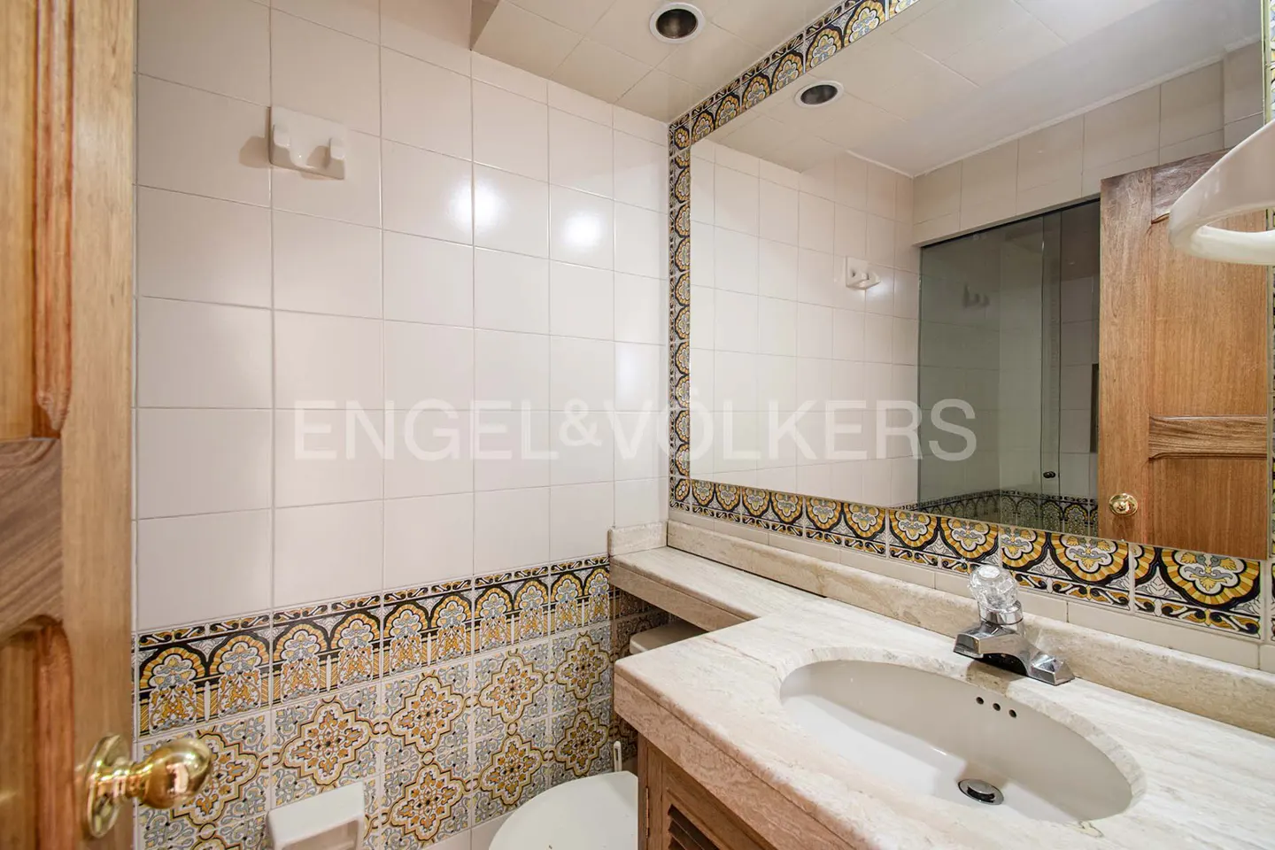 Bathroom with white tile walls, decorative tile border, marble countertop, and wood vanity. A large mirror reflects the shower.