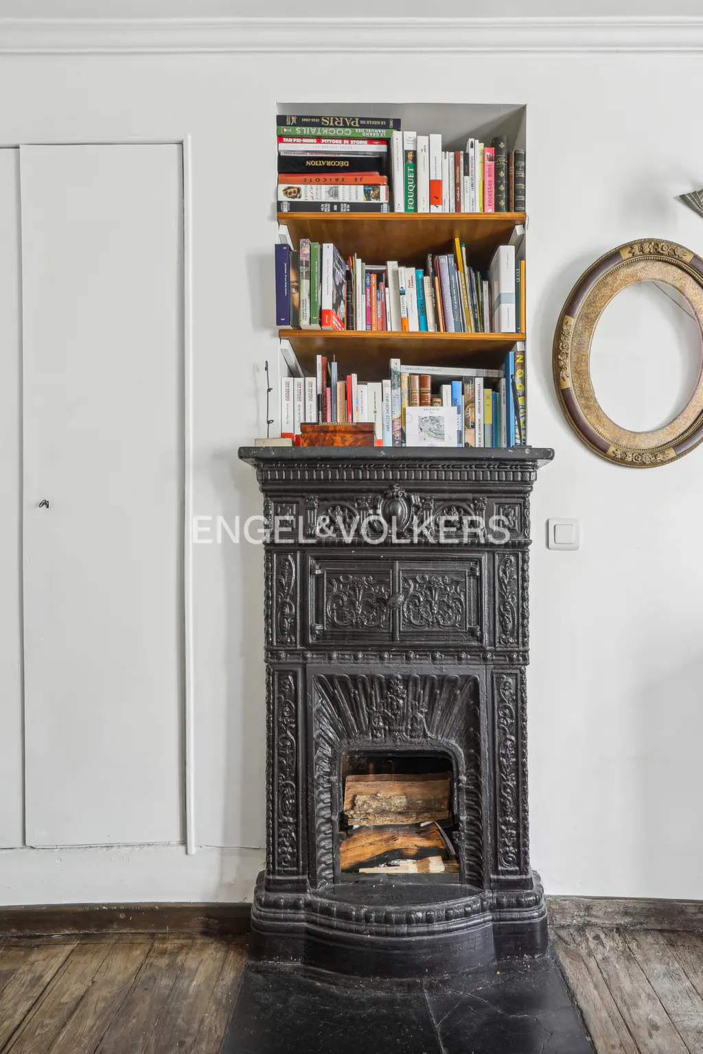 Ornate black fireplace with wood inside, built-in bookshelf above filled with colorful books, and a gold oval frame on a white wall.