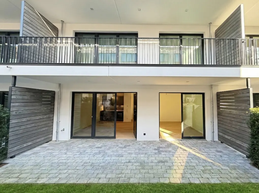 Two-story white building with black-framed sliding glass doors and a gray stone patio. Balcony with black railings on the second floor.