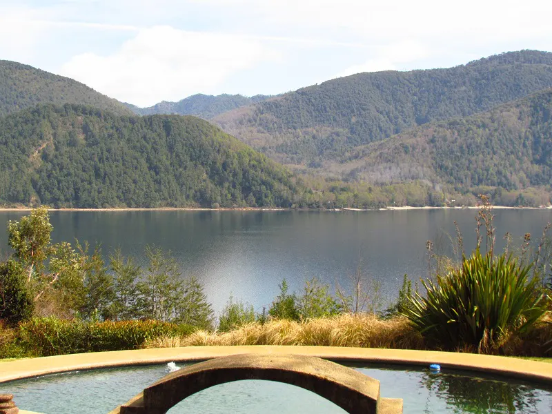 A scenic view of a pool with a stone bridge, overlooking a lake and forested mountains under a blue sky.