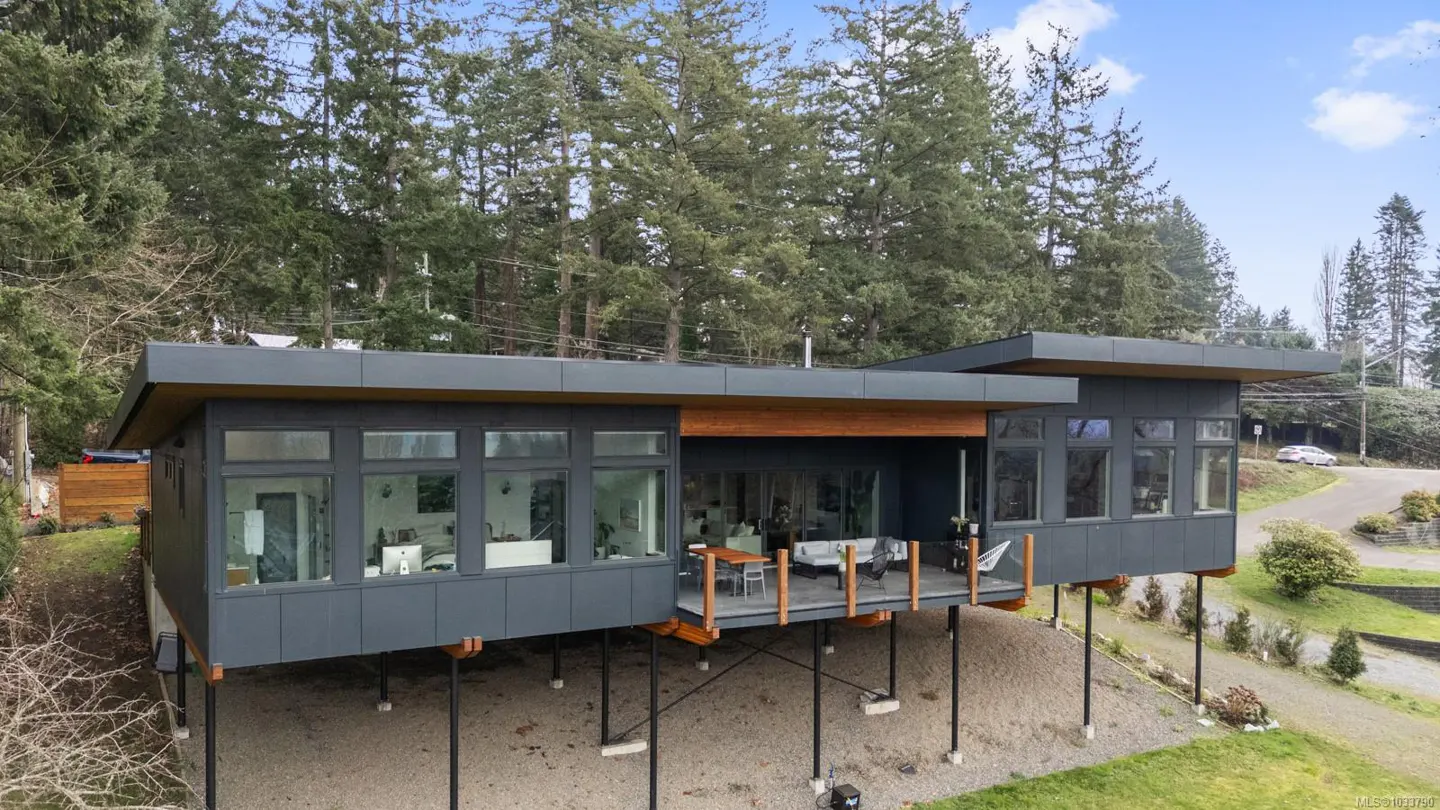 Modern gray house on stilts. Large windows, wood deck with furniture. Trees in background.