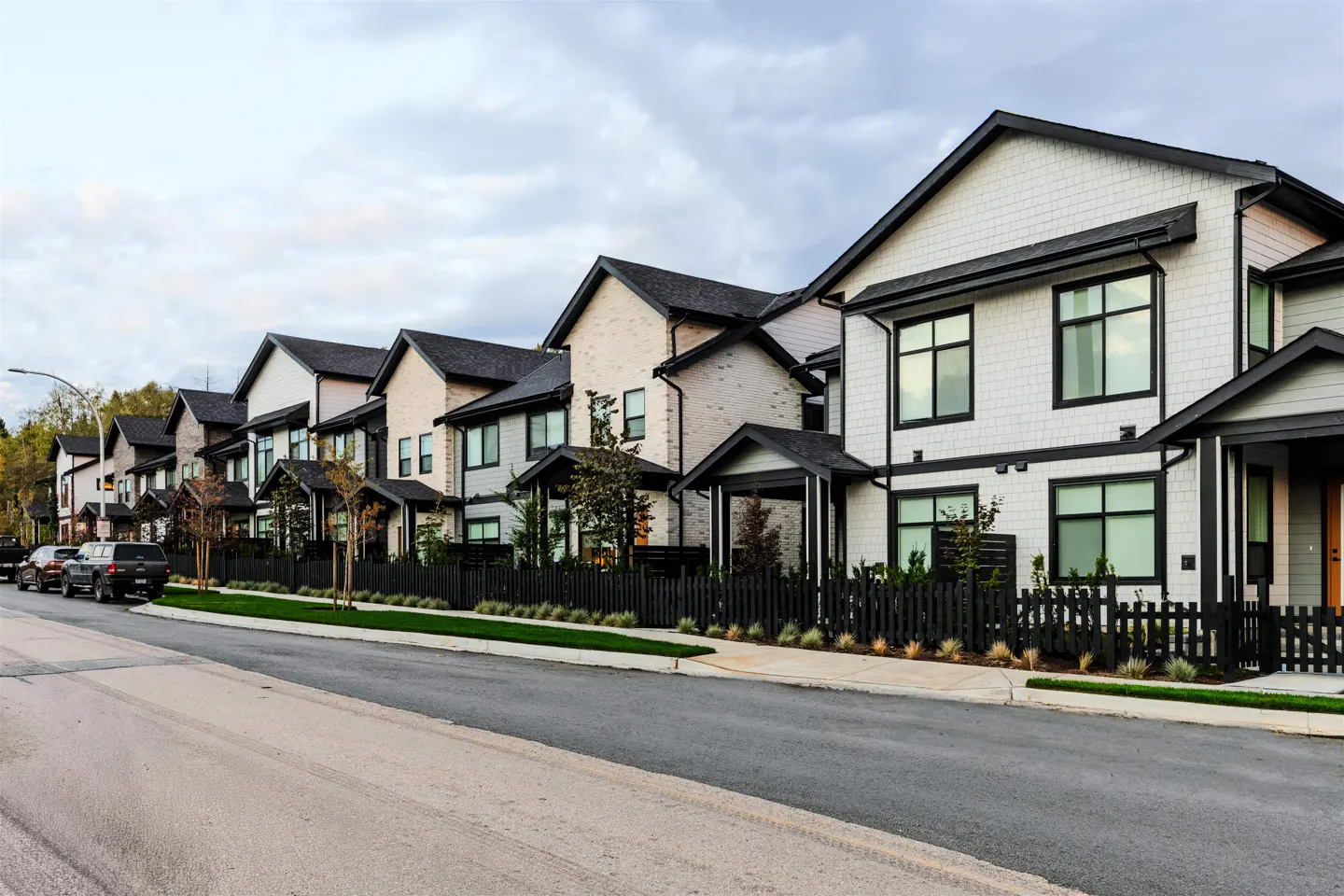 Row of modern townhouses with black trim, fences, and dark roofs, set against a cloudy sky. Cars parked along the street.