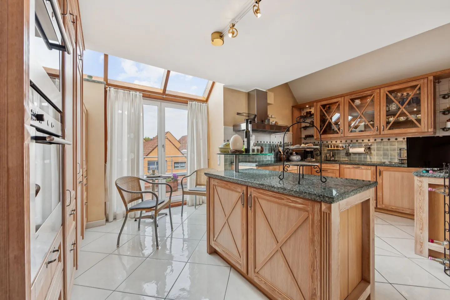 Bright kitchen with wood cabinets, granite island, and white tile floor. A table and chairs sit near a window with white curtains.