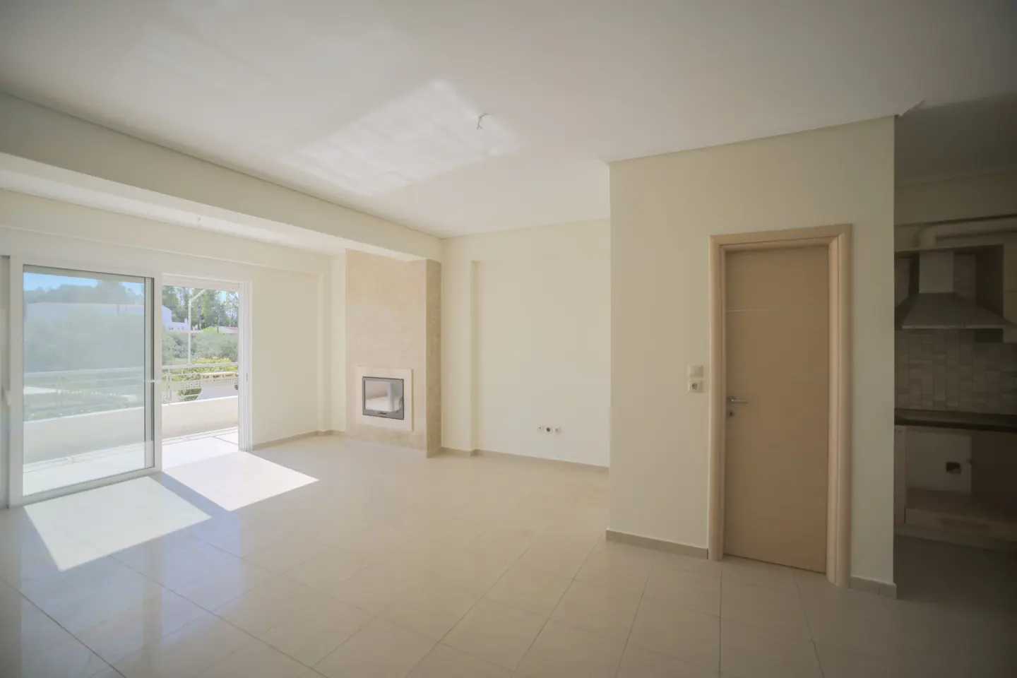 Bright, empty living room with beige walls and tile floor. Sliding glass doors lead to a balcony. A fireplace and doorway to the kitchen are visible.