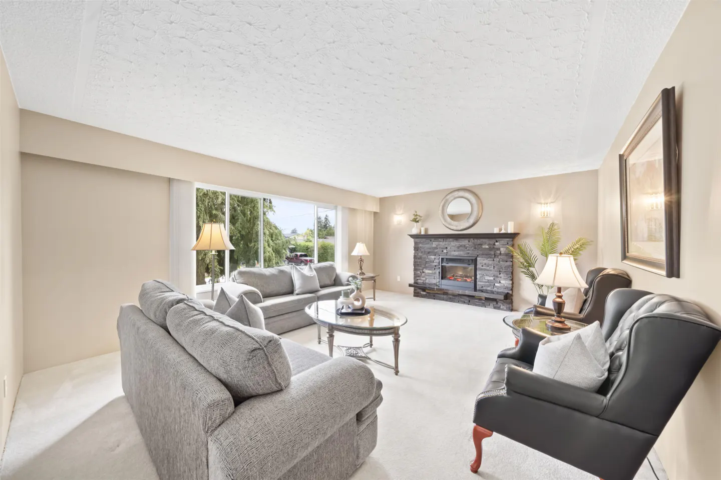 Living room with gray sofas, black armchair, stone fireplace, and large window. Beige walls and white carpet.