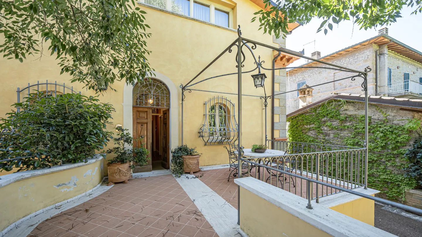 Exterior view of a yellow building with a patio, wrought iron canopy, table, and chairs. Green foliage and a stone building are visible.
