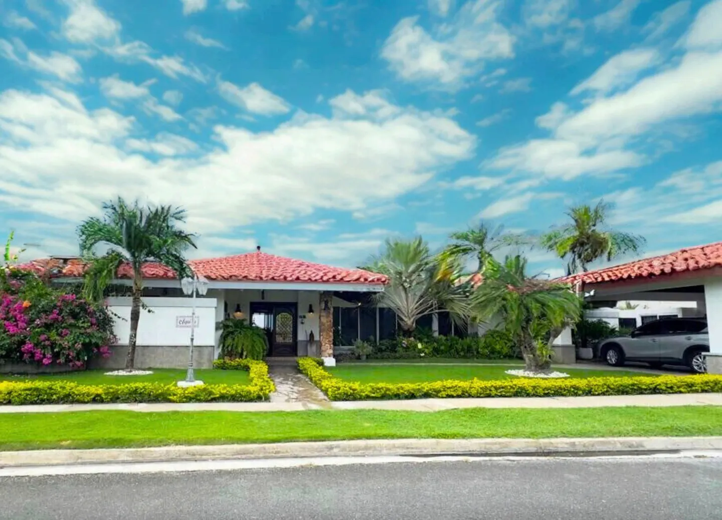 A white house with a red tile roof, green lawn, palm trees, and a blue sky with white clouds.