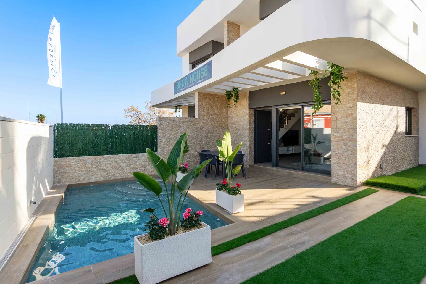 Modern home exterior with a pool, stone wall, and patio. Planters with pink flowers add color. Blue sky above.