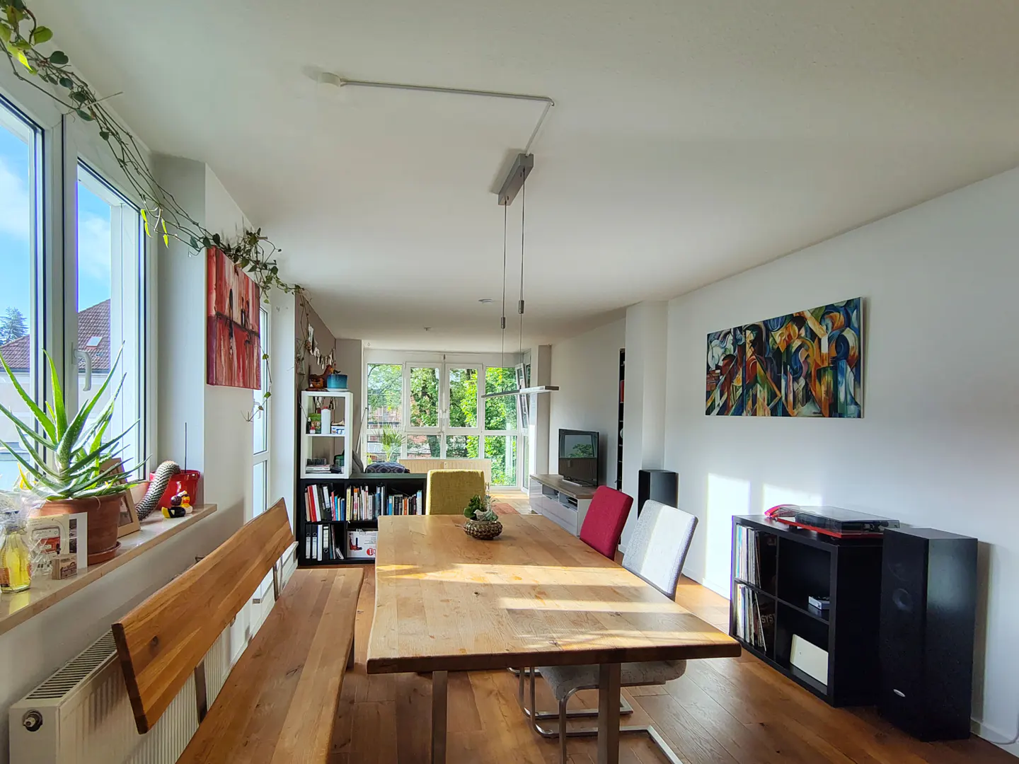 Bright, modern dining room with a long wooden table, bench seating, and large windows. Artwork and plants add color.