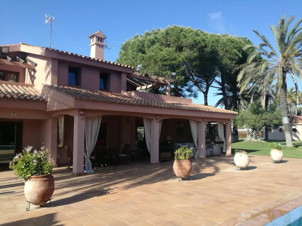 Exterior view of a salmon-colored house with a tiled roof, a covered patio with white curtains, and potted plants on a brick patio.