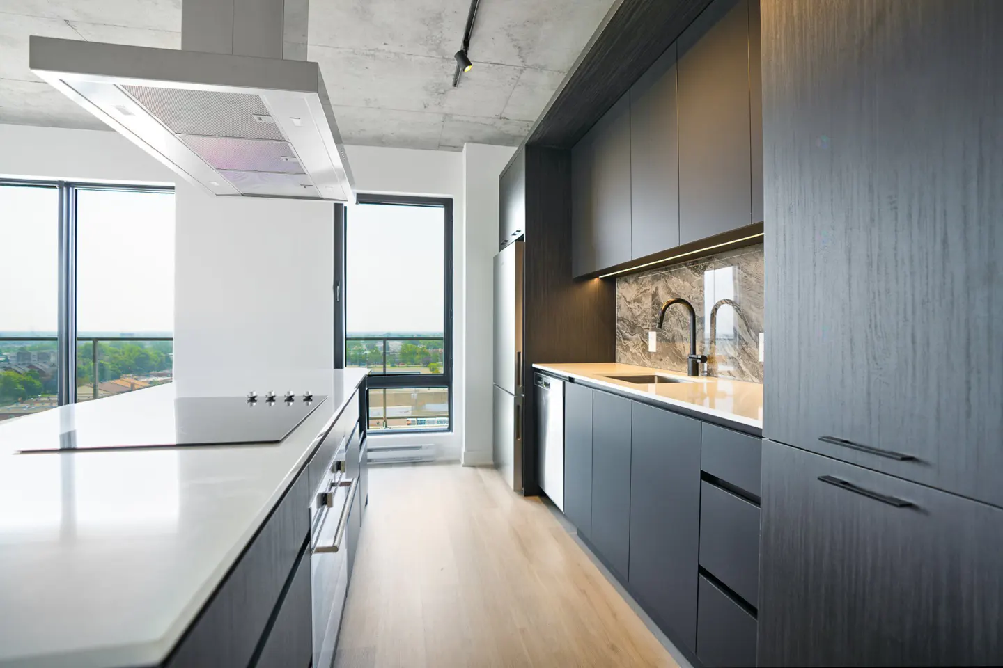 Modern kitchen with dark wood cabinets, white countertops, marble backsplash, and a concrete ceiling with large windows.