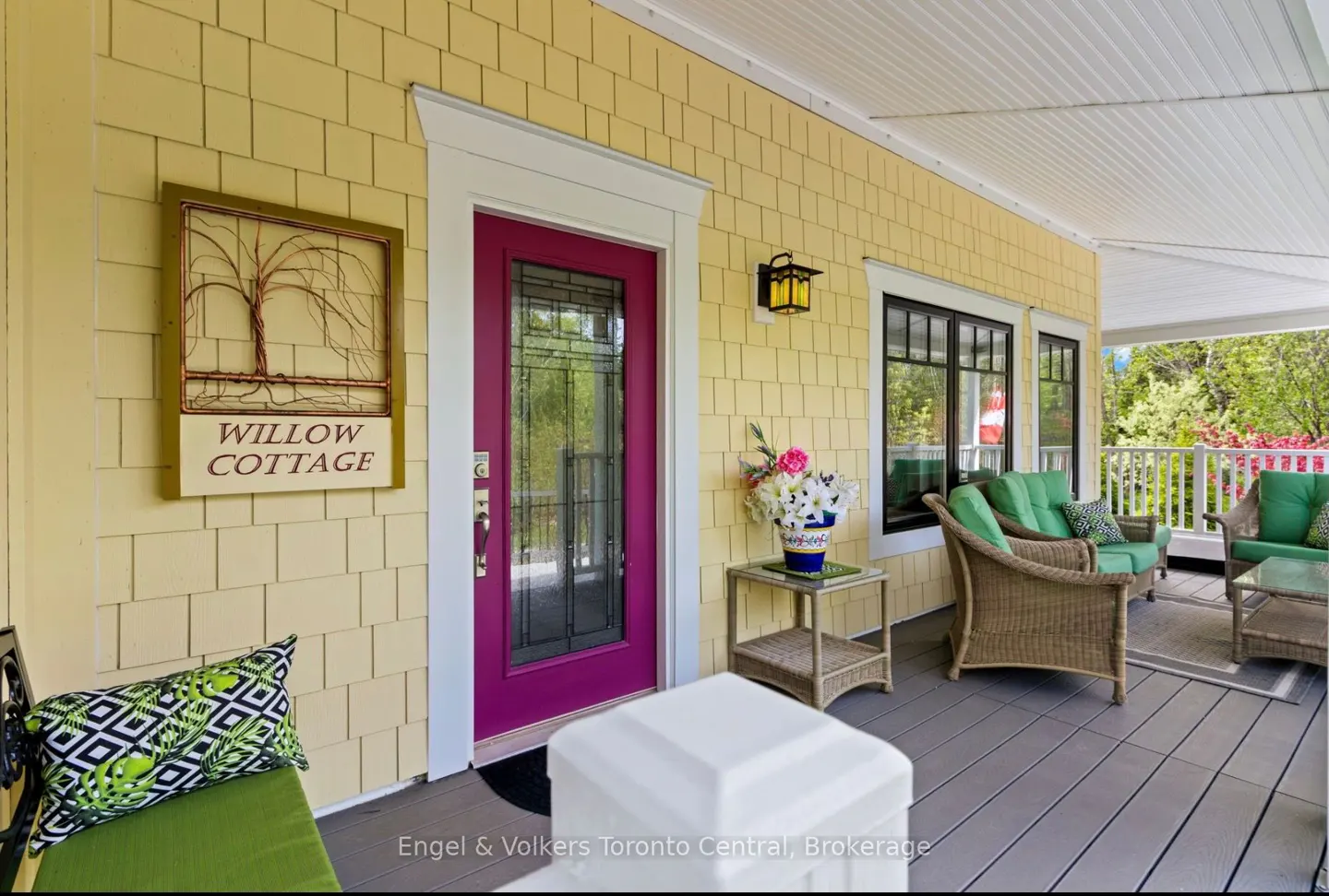 Covered porch of "Willow Cottage" with yellow siding, a magenta door, and wicker furniture with green cushions.