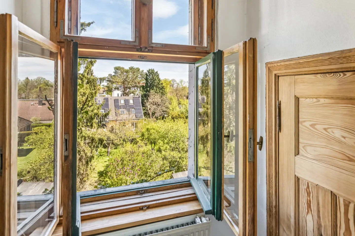 Open wooden window with a view of green trees and houses. A wooden door is visible on the right.