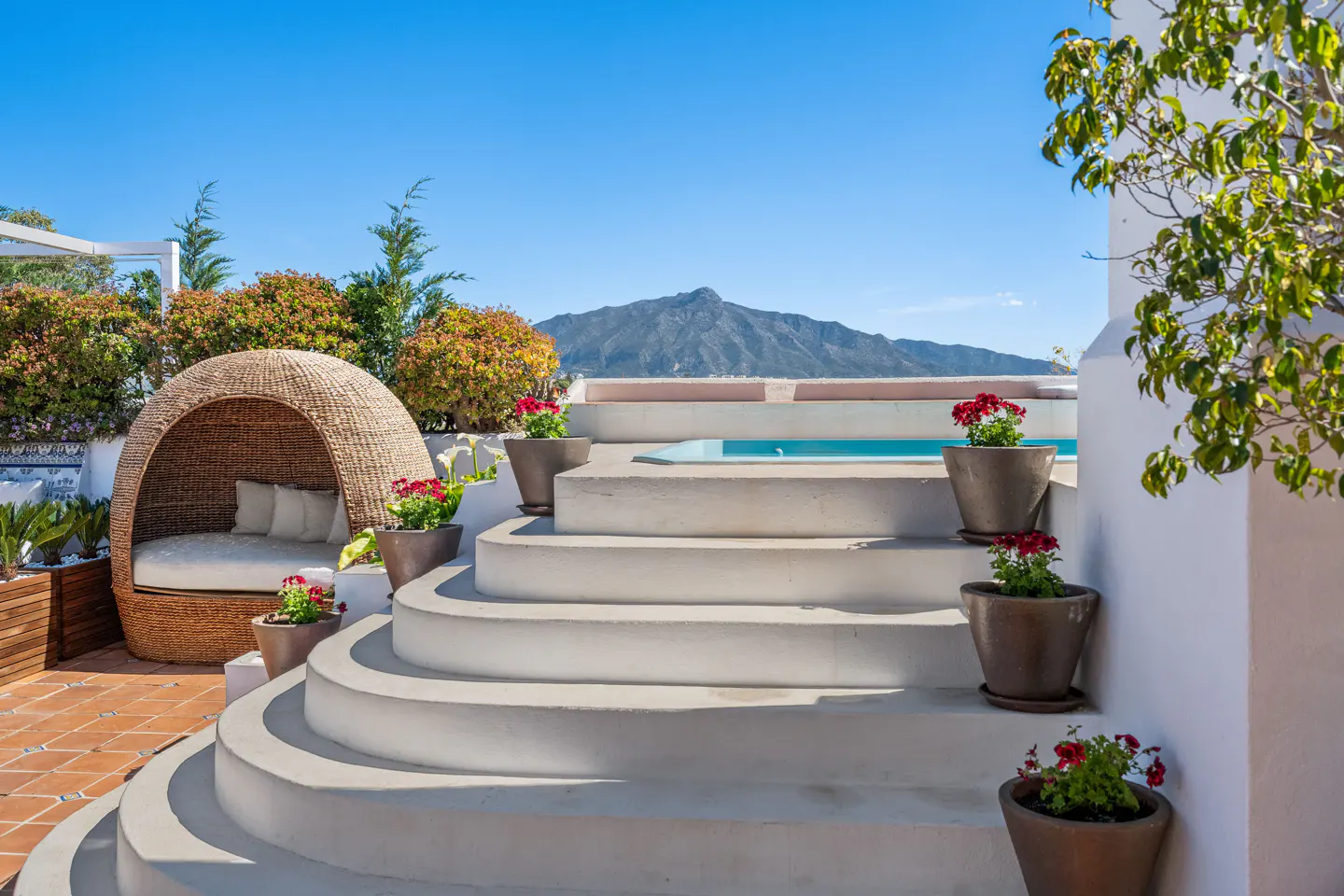 Outdoor patio with a wicker pod chair, white steps leading to a pool, potted red flowers, and a mountain view.