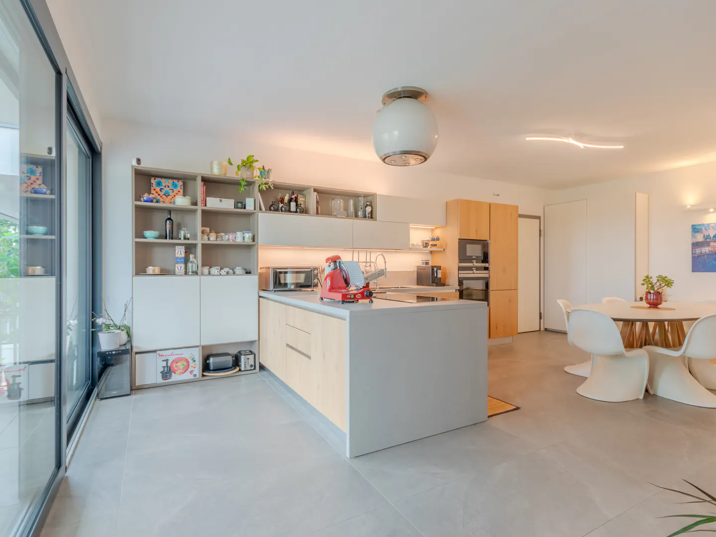 Bright, modern kitchen with an island, light wood cabinets, and open shelving. A dining area with a round table and white chairs is visible. Large windows let in natural light.