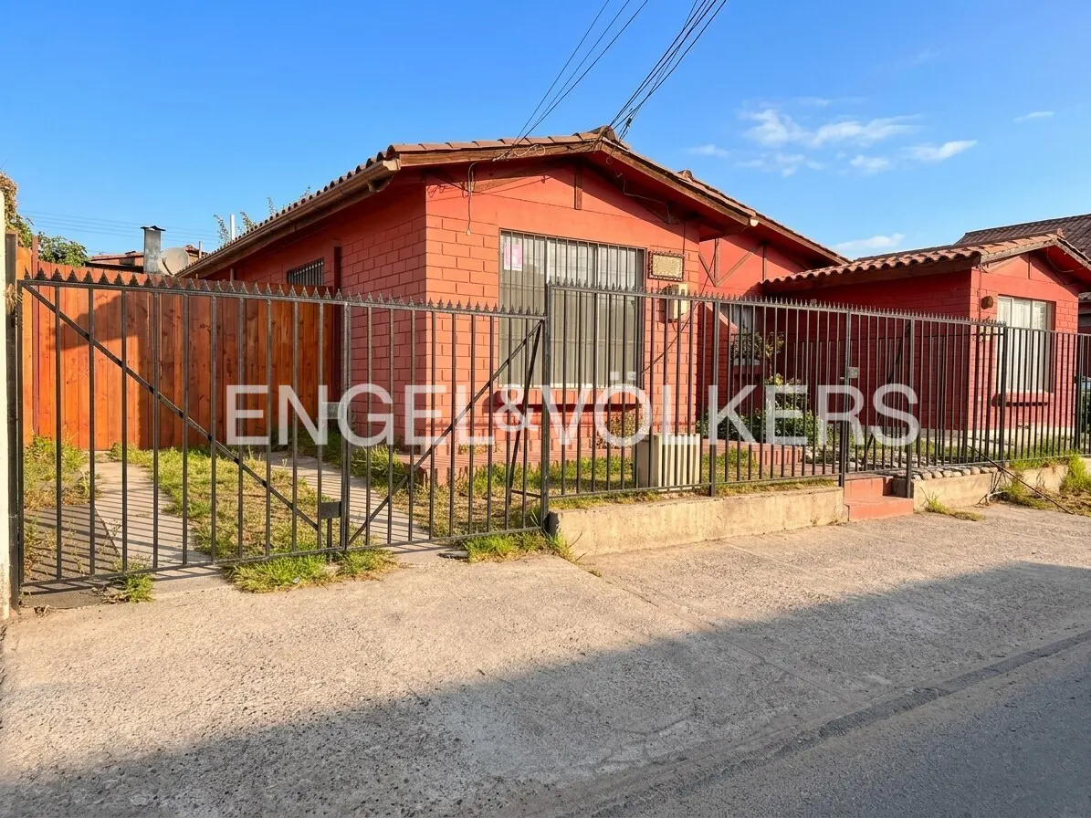 Red brick house with a brown roof behind a black metal fence and a brown wooden fence on a sunny day.