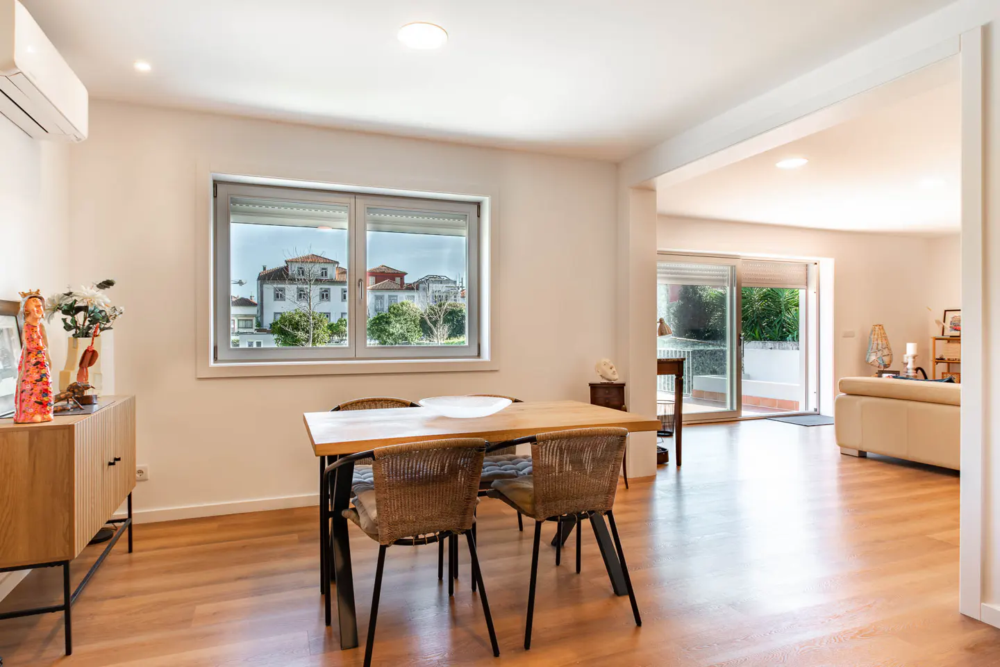 Bright dining room with wood floors, table, and wicker chairs. A window shows a view of buildings and trees. Living room visible in background.