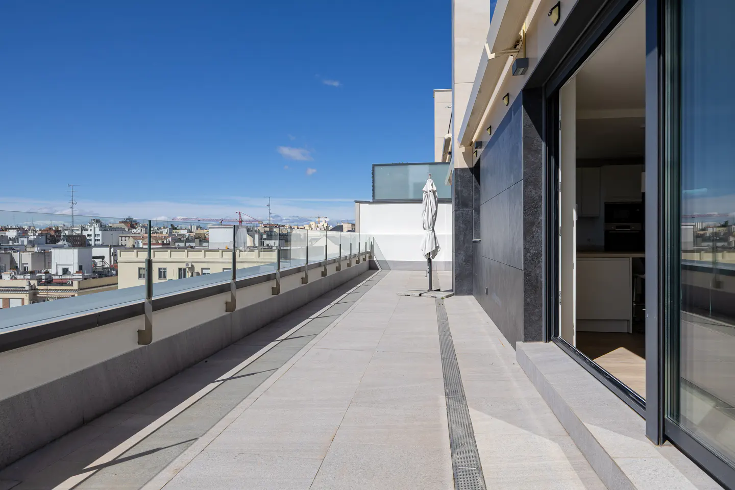A long, gray balcony with a glass railing overlooks a city under a blue sky. A closed white umbrella stands near an open sliding glass door.