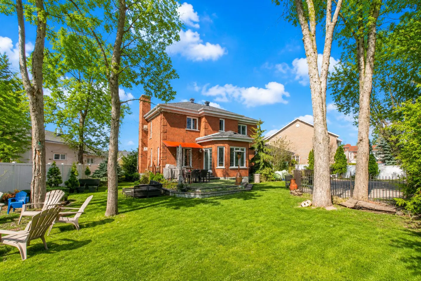 Backyard view of a two-story red brick house with a patio, green lawn, trees, and blue sky with white clouds.