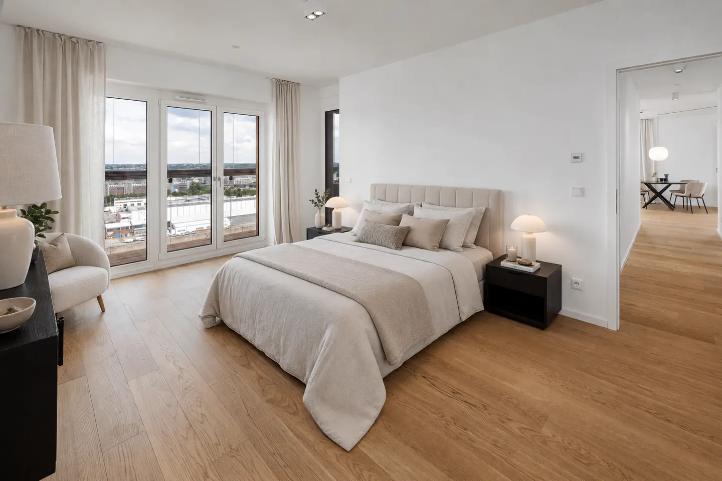 Bright bedroom with a beige bed, white walls, and light wood floors. Large windows offer a city view. A white chair sits near a black cabinet.