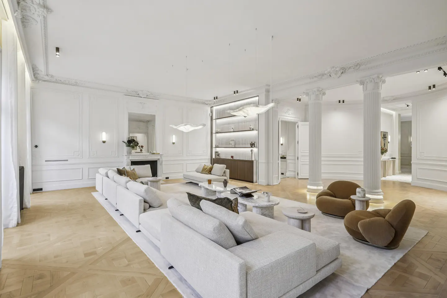 Bright, spacious living room with white walls, columns, and herringbone wood floors. Modern sofas, chairs, and tables furnish the space.