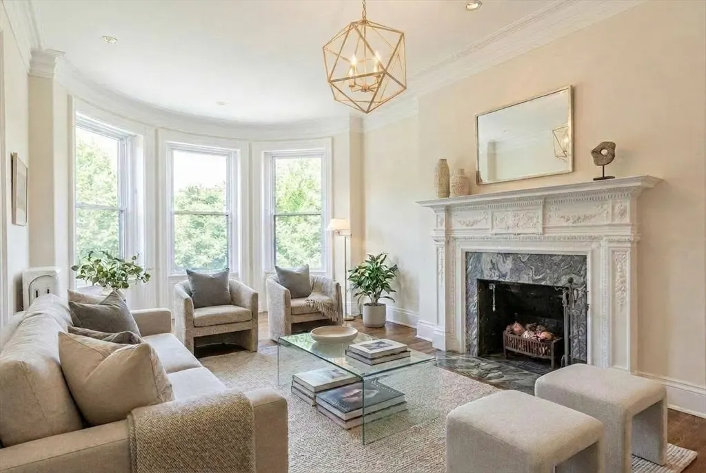 Bright living room with beige sofa, chairs, and stools. A glass coffee table sits on a patterned rug. A white fireplace and geometric chandelier add elegance.