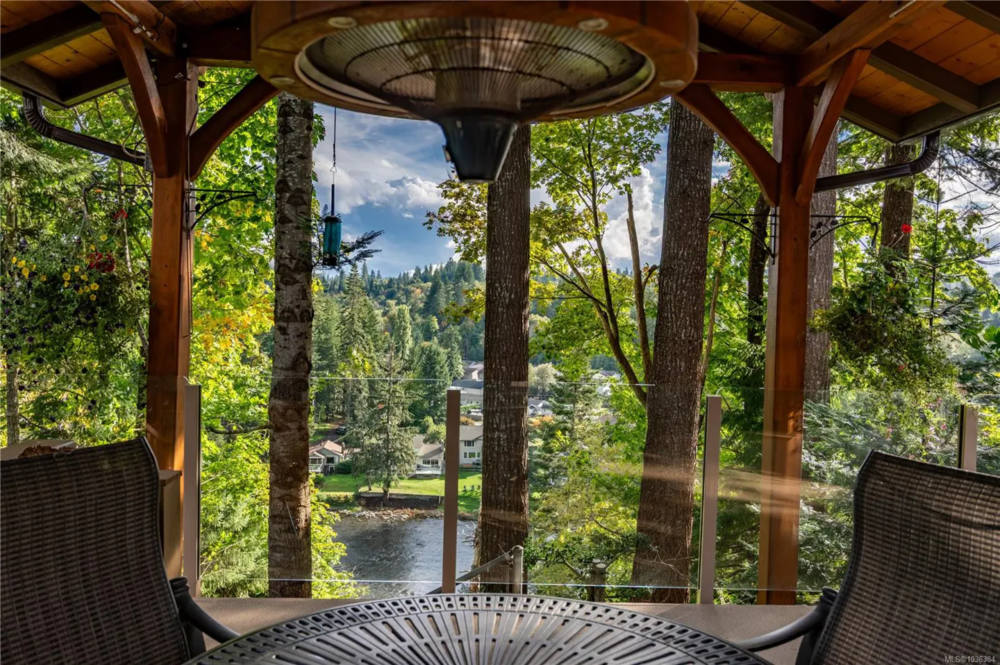 View from a wooden gazebo with a table and chairs, overlooking a lake surrounded by green trees and houses.