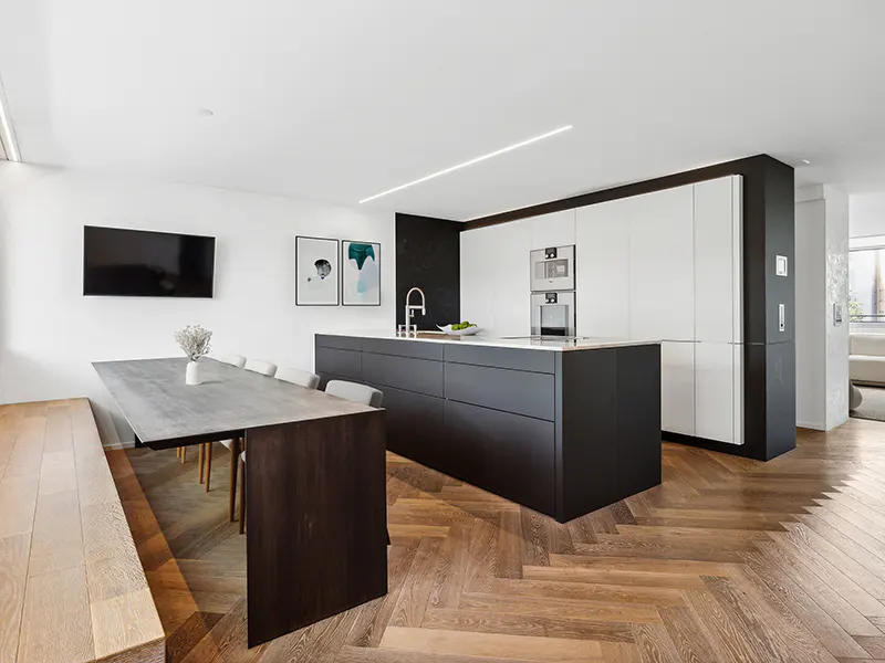 A modern kitchen with a black island, white cabinets, and a wooden herringbone floor. A dining table with a bench is to the left.