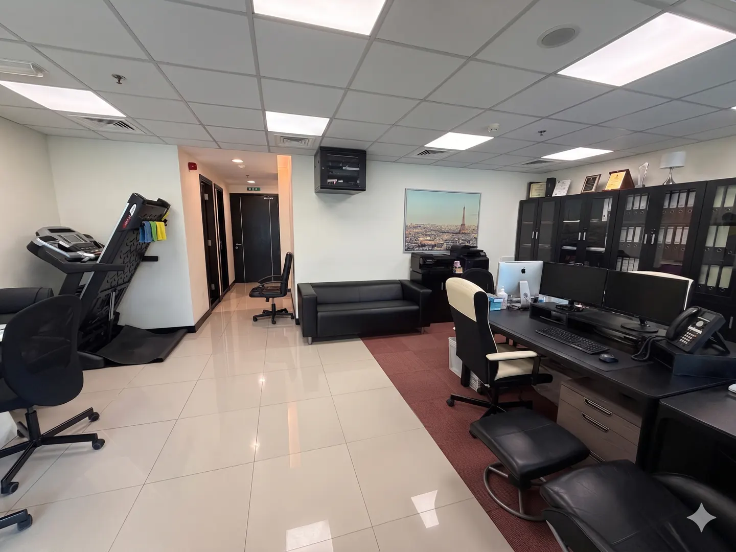 Office space with a treadmill, black sofa, and desk with computer monitors. A picture of Paris hangs on the wall.